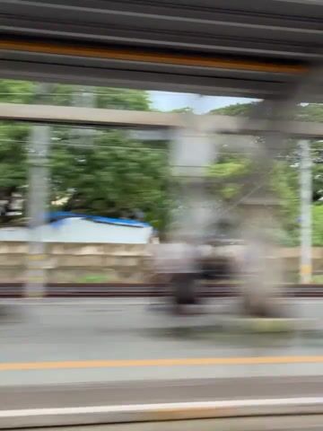 The video shows a blurry view from a moving vehicle, likely a train, passing a station. The scene is characterized by motion blur, making specific details difficult to discern. There are platforms with yellow lines, overhead structures, and buildings in the background. Some indistinct figures, presumably people, can be seen on the platform, as well as what appear to be shops or kiosks. The overall impression is of a busy urban environment viewed from a rapid transit system.