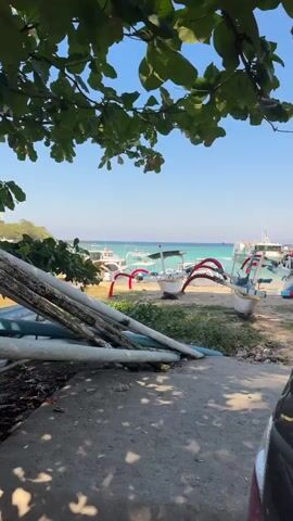 The video shows a view of a beach with several boats parked on the sand and in the shallow water. The boats appear to be traditional fishing boats, with outriggers and a canopy. The water is a clear turquoise color, and the sky is a bright blue with no clouds. Lush green trees with dark green leaves hang over the top of the frame, casting shadows on the ground. The ground in the foreground is a mix of sand and concrete, with some debris and what appears to be a black car bumper on the right side of the frame. The overall scene is peaceful and tropical, suggesting a sunny day at the beach.