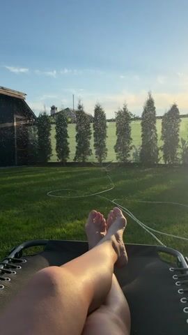 This media file captures a serene moment in a backyard setting in Kurylivka, Ukraine. The subject is a person, seen from the knees down, relaxing on a lounge chair. Their legs are crossed, and their feet are visible, with painted toenails.

The scene is a lush green lawn, with a row of tall, slender trees forming a natural border along the back of the yard, behind a fence. A garden sprinkler is active, casting a fine mist of water into the air, creating a shimmering effect. The sunlight suggests it is either early morning or late afternoon, with a warm glow illuminating the scene. The sky is a clear blue, with a few wispy clouds, indicating pleasant weather.

A dark-colored shed or outbuilding is partially visible on the left side of the frame, providing a hint of the domestic setting. The overall mood is one of relaxation and enjoyment of a peaceful outdoor environment. There is no visible text in the media file.