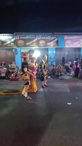 The video shows a street scene at night with people gathered. In the foreground, a few individuals in traditional costumes are standing in the street. Behind them, a crowd of spectators is seated on the ground, watching. A motorcycle is seen moving across the frame. The background features a building with illuminated signage. The lighting suggests an outdoor evening event.