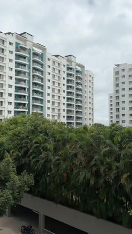 The video shows a medium shot panning across two modern, white apartment buildings with balconies and teal accents. In front of the buildings, there is a dense line of lush green trees and foliage. The sky is overcast with clouds. At the bottom left of the frame, a portion of a dark gray structure, possibly a covered walkway or parking area, is visible, along with the front end of a black motorcycle. The camera movement is smooth and slow, creating a gentle sweep from left to right.