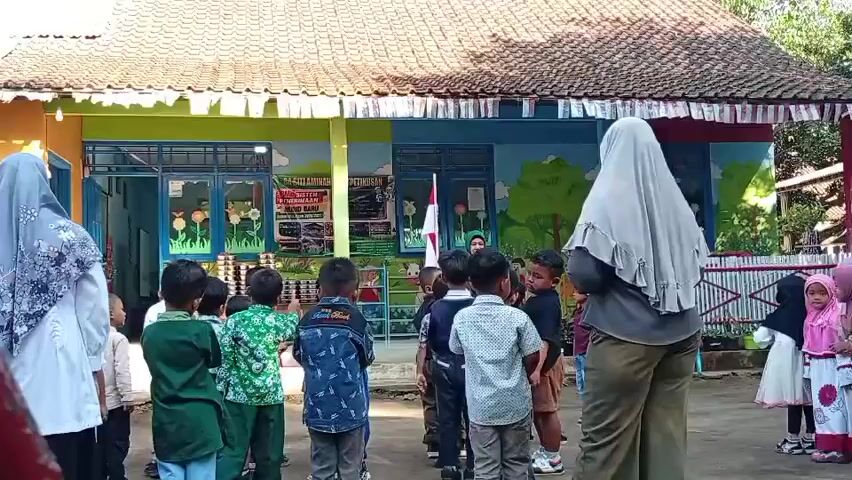 In front of a brightly colored, single-story building with a tiled roof, a group of young children are gathered outside. Some of the children are wearing uniforms, including some in green and blue. Two adults, likely teachers, are also present, one on the far left wearing a light-colored hijab and tunic, and another on the right wearing a gray hijab and olive-colored pants. The building appears to be a school or daycare, with colorful murals of cartoon landscapes on its exterior walls. The children are standing in lines, facing towards the building, and some appear to be listening to instructions from one of the adults. A flagpole with a flag is visible in front of the building. The ground is paved, and there are some plants and fences visible in the background. The scene appears to be a school assembly or outdoor activity.