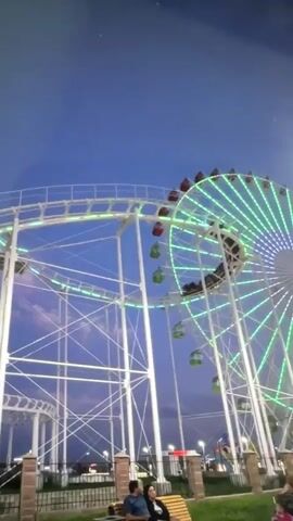 A vertical, somewhat blurry, low-light video captures an amusement park scene at dusk or night. Dominating the frame are the tracks of a white roller coaster, illuminated by green and white lights, winding upwards and then descending. To the right, a Ferris wheel, also lit with green lights, is visible. In the foreground, two people are seated on a wooden bench, facing away from the camera. They appear to be a man and a woman. The sky is a deep blue with some wispy clouds. Green grass and a brick fence are also visible in the lower part of the frame. A roller coaster car filled with passengers can be seen moving along the track in the upper portion of the video. The overall atmosphere suggests a lively, illuminated evening at an amusement park.