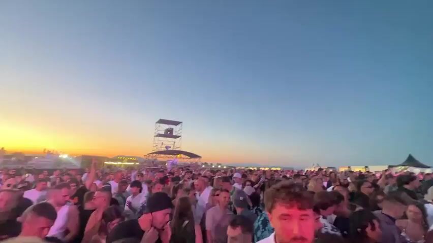 A wide shot of a large outdoor gathering of people. The sky is clear and blue, with the sun setting on the horizon, casting a warm orange glow. In the background, a large stage structure with speakers and lights is visible. There are also tents and food vendors set up. The crowd is dense and appears to be enjoying themselves, with many people dancing and socializing. The camera pans across the crowd, showing a variety of faces and expressions. The overall atmosphere is festive and energetic.