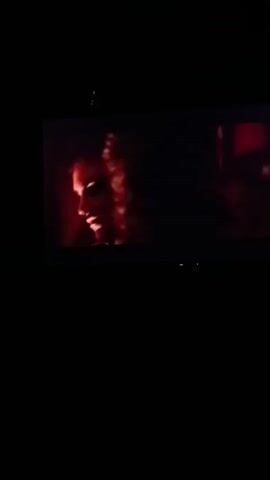 A dark, dimly lit room features a large television screen displaying a close-up of a man's face. The man has dark, curly hair and is looking down with a serious expression. He is wearing a white uniform with gold trim. The lighting on the screen is warm and dramatic, with a red hue. The room itself is entirely black except for the glow of the television.