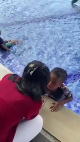 The video shows children in a large, outdoor swimming pool with blue tile. The pool has a shallow area where some kids are sitting and splashing. Other children are running through the water. There is a play structure in the pool with two slides. The background shows a green, hilly landscape with some structures. A Ferris wheel is visible in the distance. A woman in a red shirt is seen kneeling at the edge of the pool, interacting with a child.