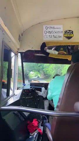 The video, shot from inside a vehicle, shows a person wearing a light green shirt and white gloves, operating the steering wheel and controls of what appears to be a bus or large truck. The driver is leaning forward, facing the dashboard which is equipped with numerous buttons and switches. The vehicle is on a road with trees and greenery visible outside the windshield. The driver's right hand is holding a white cloth or glove. A white piece of paper with handwritten numbers and text is affixed to the ceiling above the driver's area. A yellow and brown plush headrest or pillow is also visible. The overall environment suggests an outdoor journey.