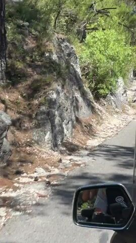 This is a first-person perspective video from a motorcycle or scooter traveling on a paved road alongside a rocky, wooded hillside. The road is on the right side of the frame, curving slightly to the right. A motorcycle's windshield and side mirror are visible in the foreground, reflecting the rider and the road behind them. The hillside is covered with trees and shrubs, and some rock formations are visible. Sunlight is dappling through the trees, indicating it is daytime. In the distance, other vehicles can be seen on the road, and there appear to be some signs. The video is shaky, suggesting the vehicle is in motion.