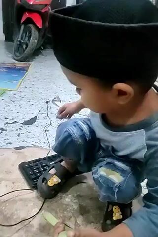 A boy wearing a grey shirt and blue shorts is squatting on the floor, holding a green object in his left hand and a keyboard in front of him. Another person, only their legs and torso are visible, wearing shorts and holding a smartphone, is standing over the boy. The smartphone is positioned above the boy's head, displaying an image or video. The environment appears to be indoors, with a tiled floor and various objects in the background, including a fan, a red toy vehicle, and a play mat with numbers and animals. The lighting suggests it is daytime.