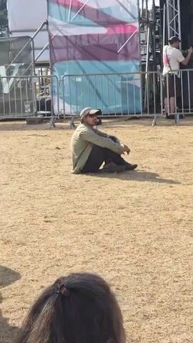 A man wearing a hat and sunglasses sits on the ground. He is wearing a light green jacket over a dark shirt and dark pants. He has dark shoes. He is facing to the left, looking away from the camera. Behind him is a large white structure with pink and purple stripes, and a metal fence. A person is visible to the right of the frame, wearing a white shirt and shorts, appearing to be working on the structure. In the foreground, the top of another person's head is visible, with brown hair. The ground is covered in dry grass and dirt.