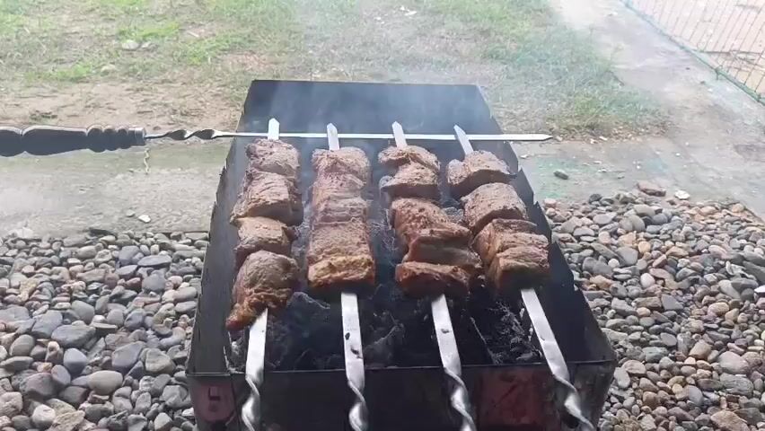 Four skewers of meat are being grilled over hot coals in a black metal barbecue. The meat is cut into large cubes and appears to be cooking evenly, with some charring visible. Smoke rises from the barbecue, indicating active cooking. The barbecue is placed on a bed of small, smooth stones. In the background, there is a grassy area and a fence. The overall scene depicts an outdoor cooking setup.