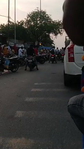 The video shows a busy street scene with many motorcycles parked on the side of the road. People are walking around and interacting, some are on motorcycles. There are buildings and trees in the background. The lighting suggests it is late afternoon or early evening. The overall impression is one of a bustling marketplace or a busy urban area.