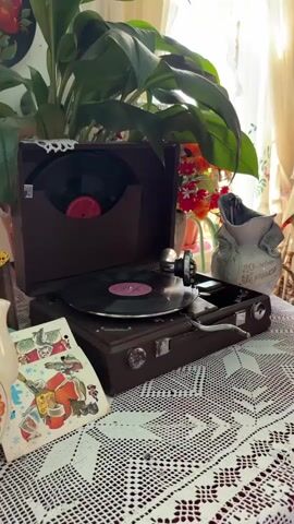A vintage record player is shown on top of a white crocheted tablecloth. A record is spinning on the player. Behind it is a tall potted plant with large leaves. A vase with flowers is next to the plant. A few vintage cards are visible next to the record player. The entire scene has a vintage and cozy aesthetic.