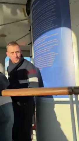 Inside a glass-walled structure, likely a rotating viewing platform, several people are visible. A man wearing a dark jacket with white stripes is standing near a wooden railing, looking out.  Behind him is a cylindrical structure with a blue informational poster attached to it. Next to the poster, a small niche contains a red fire extinguisher. A man in a black uniform with "STAFF" printed on the back is standing near the window, seemingly attending to something. The overall environment appears bright and the scene is well-lit.