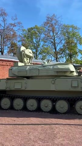 This video shows a row of military vehicles parked outdoors on a sunny day. The vehicles are painted a camouflage green and appear to be in a museum or exhibition setting. The ground is covered in red gravel, and a paved walkway runs along the side. In the background, there are trees with green foliage and a brick building. A flag with a ship and maritime symbols is visible on a flagpole to the left of the frame. The sky is blue with a few white clouds. The camera pans from left to right, showcasing several tracked and wheeled vehicles, suggesting a collection of military hardware. The overall mood is calm and observational, with no people visible in the frame. The time of day appears to be daytime, likely late morning or afternoon, given the brightness of the sun and the shadows cast. The location is identified as Kronstadt, Russia. No visible text is discernible on the vehicles or surroundings.