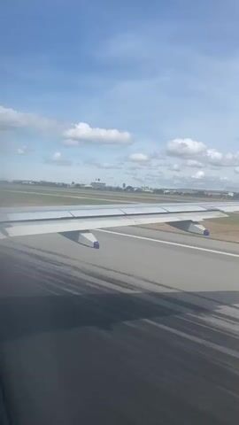 The view from an airplane window shows the plane speeding down a runway, preparing for takeoff. The wing of the plane is prominently displayed in the foreground, with the runway stretching out in front. There are white lines painted on the gray runway. Beyond the runway is a grassy area, and in the distance, there are buildings and structures suggesting an airport complex. The sky is blue with scattered white clouds. The lighting suggests it is daytime. The camera appears stationary within the plane, capturing the movement of the ground as the plane accelerates.