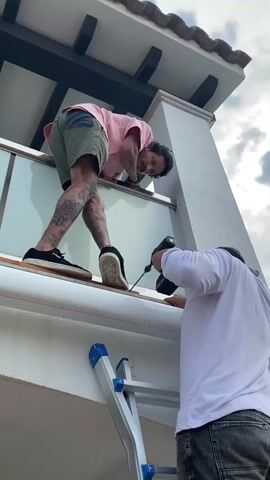 The video shows two men working on the exterior of a building. One man, wearing a pink t-shirt and green shorts, is leaning over a railing on an upper level, holding onto the railing with his left hand and looking down. He has tattoos on his arms and legs. The other man, wearing a white long-sleeved shirt and dark pants, is standing on a silver ladder and using a black cordless drill to screw something into a white surface below the railing. The background shows a cloudy sky and a white pillar.