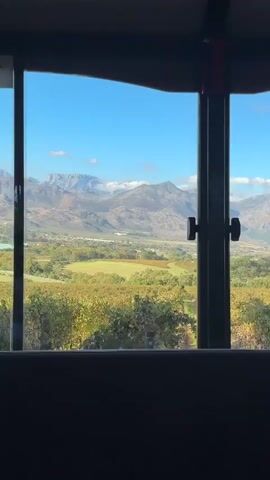 A static, interior shot shows a window framing a landscape view. In the foreground, a vineyard with rows of green plants. Beyond the vineyard is a valley dotted with green fields and buildings. In the distance, mountains loom under a clear blue sky with scattered white clouds. The window frame is black, and the interior of the room is dark. The scene has a peaceful and scenic quality.