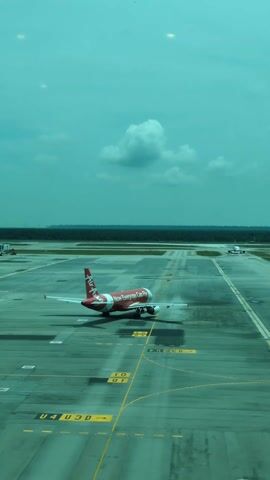 The video shows a red and white Air Asia plane standing on a runway at an airport. Another plane is visible in the distance. There is a large forest behind the runway. The sky is turquoise. The view is from inside an airport building, looking out through glass. Markings and painted lines are visible on the runway, and arrows painted on the ground point in various directions.
