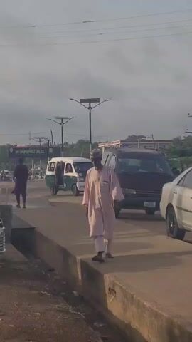 A man wearing a light pink traditional outfit and a cap walks along the side of a road. The road is lined with cars and there is a pharmacy sign in the background. The man is walking from right to left and appears to be the main subject of the video. He walks into the distance.