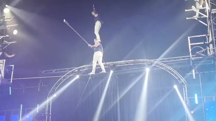 This media file captures a thrilling circus performance. Two male acrobats are the central subjects, engaged in a high-wire balancing act. One acrobat stands atop the other's shoulders, while the bottom acrobat skillfully balances on a tightrope stretched across an elevated arched structure. The bottom acrobat also holds a long pole, extending it forward and likely using it for balance. Several other performers are visible on elevated platforms to the left and right, some appearing to be on bicycles, suggesting a multi-faceted performance. The setting is a dimly lit indoor arena, illuminated by dramatic stage lighting, with bright white spotlights creating beams that cut through the haze. The overall atmosphere is one of suspense and daring. The time of day is not discernible, but the indoor setting and artificial lighting indicate a performance taking place in the evening or night. There is no visible text within the frame.