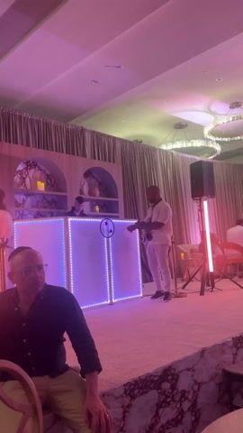 The video shows a brightly lit indoor event space with a DJ booth as the main focal point. A man in a white outfit stands to the side of the booth, interacting with the DJ, who is positioned behind the console. A woman with long dark hair stands nearby. The DJ booth is illuminated with purple and blue LED lights, and its front features a white panel with a black and white logo. To the left of the booth is a seated man with glasses, who is looking towards the DJ. The room has a low ceiling, chandeliers, and beige curtains lining the back. There are decorative shelves on the wall behind the booth. A speaker stands on a tripod to the right, lit by a red light. A security rope is in place to keep the crowd at bay. The event has soft lighting throughout.