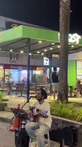 A man in a white t-shirt and blue jeans sits on the ground playing an acoustic guitar. He is wearing a black mask. A microphone is positioned in front of him. He is surrounded by palm trees and a building with green and white accents. The building has signs for "Boost" and "Dessert." There are lights strung across the area. The scene appears to be outdoors at night. Other people can be seen in the background, sitting at tables outside the building.