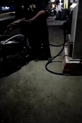 The video shows a person wearing a dark shirt and pants, and a yellow baseball cap, standing next to a motorcycle at what appears to be a gas station at night. The person is holding a fuel nozzle and is in the process of refueling the motorcycle. The gas pump is visible to the right of the frame. The ground is paved and appears to be wet. Other motorcycles and some structures are visible in the background, illuminated by artificial lighting. The scene is dimly lit, suggesting it is nighttime.