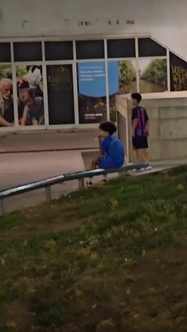 The video shows a group of children playing with a soccer ball outside at night. In the foreground, a metal railing runs along a grassy slope. In the background, a building with large windows displays images of people, possibly for advertising. Two young boys are visible, one wearing a blue shirt and the other a purple and blue striped shirt. The boy in the blue shirt is sitting on the railing, while the boy in the striped shirt is running and kicking a soccer ball. Another child is blurred in the background to the right. The lighting suggests it is nighttime, with artificial lights illuminating the scene.
