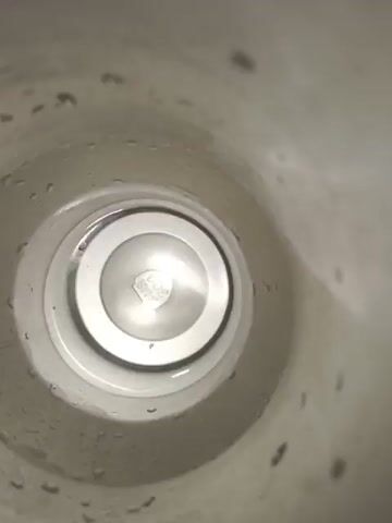 The video is a first-person view from inside a container, possibly a bottle or a cup, looking down into its opening. The walls of the container are light-colored and appear to have water droplets clinging to them. The bottom of the container is visible, with a small amount of clear liquid at the very bottom. The lighting is bright, creating reflections on the liquid and the inner surface of the container. The perspective suggests that the viewer is looking down into the container from the top. There are no people or other objects visible in the video. The scene is static, with no apparent movement.