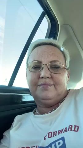 The video shows a close-up selfie of a middle-aged white woman with short blonde hair and glasses. She is wearing a white t-shirt with red lettering and a beaded necklace. The woman is inside a vehicle, as indicated by the window and car interior visible in the background. The lighting suggests it is daytime, and the sky is visible through the window. The woman looks at the camera and smiles.