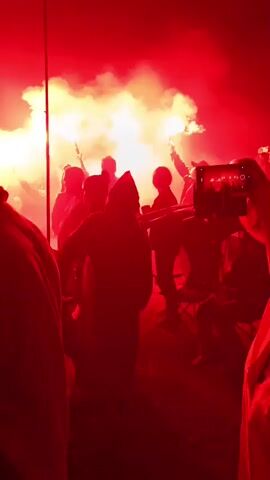 A group of people are silhouetted against bright red flares. One person in the foreground is holding a smartphone to record the scene. Several individuals are wearing hooded cloaks. The environment appears to be outdoors at night.