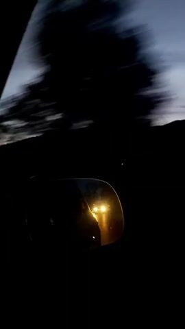 The video shows a rearview mirror reflecting a bright light as a vehicle travels. The sky in the background appears to be twilight, with hills or mountains silhouetted against the pale sky. Trees or brush are visible in the periphery, blurred due to motion. The overall impression is of a journey taking place at dusk or dawn.