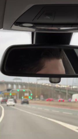 The video is shot from inside a car, looking out through the windshield and the rearview mirror. In the rearview mirror, you can see the top of a person's head. Outside, the car is driving on a multi-lane highway. There are other cars on the road, including a yellow Nissan Juke in front of the camera car. In the background, there is a bridge with two tall white towers. There are streetlights along the highway. The sky is overcast. The music is hip-hop.