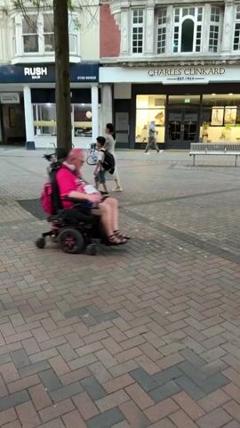 A man with pink hair is riding a black electric wheelchair on a brick-paved street in what appears to be a town or city. He's wearing a bright pink shirt, dark shorts, and sandals, and has a pink backpack on the back of the wheelchair. He's looking at a cell phone he holds in his hands. There are buildings visible in the background, some with storefronts and other people walking or sitting in the vicinity.
