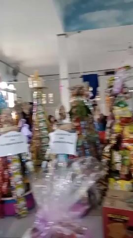 The video pans across a room that appears to be a mosque or community center, decorated with what seems to be offerings or celebratory items on shelves and the floor. In the background, several women are seated on the floor, facing away from the camera, some wearing hijabs. Further into the room, a man in traditional attire, including a skullcap, is seated on the floor facing a wooden pulpit. The room has white walls, green accents on arched doorways, and a green carpet covering the floor. There are also windows visible, letting in natural light. The overall atmosphere suggests a religious gathering or event.