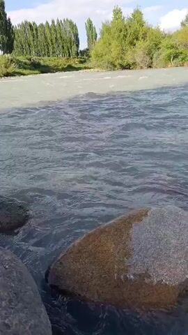 The video shows a flowing river with rocks in the foreground and along the bank. Trees and vegetation line the opposite bank, with a few scattered structures visible in the distance. The sky is partly cloudy. The camera pans slowly across the scene, capturing the movement of the water and the natural landscape.