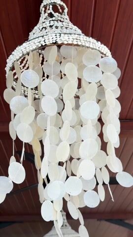 The video shows a hanging chandelier type object with numerous white mother-of-pearl disc shaped pendants attached to a white beaded frame. The structure hangs from a dark brown, plank ceiling and is being swayed. The sound of the pendants tinkling together can be heard.