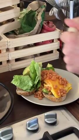 The video shows a close-up shot of a plate of food with a hand squeezing hot sauce onto it. The plate contains a sandwich with whole-grain bread, avocado slices, lettuce, scrambled eggs, and baked beans. A bottle of hot sauce is being squeezed, and the sauce is visible on the eggs and beans. The background shows a wooden crate and kitchen utensils. The camera is positioned at eye level with the plate of food.