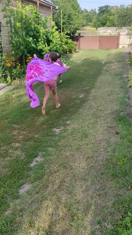 A child is spinning in a garden, holding a large purple cloth with pink flower designs. The child is wearing a pink dress and is barefoot. The garden has green grass, trees, and flowers. The child spins for several seconds, with the cloth flowing around them. The video is shot from a slightly elevated angle, looking down at the scene.