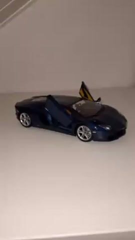 The video shows a dark blue Lamborghini Aventador model car on a white surface. The car's doors are open, revealing a yellow interior. The camera angle is slightly above the car. The background is a blurry white or light gray wall with a hint of a white shelf or structure.
