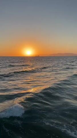 The video shows a sunset over a body of water. The sun is a bright orange disc low on the horizon, casting a golden reflection on the rippling waves. In the distance, there are faint outlines of land or islands. A large ship is visible on the horizon, silhouetted against the sunset. The sky is a gradient of pale blue at the top, transitioning to orange and yellow near the horizon. The water is a deep blue-green, with white foam cresting on some of the waves in the foreground. The overall impression is a calm and serene natural landscape.