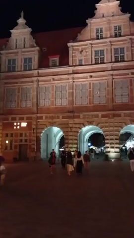 The video begins with a nighttime view of an ornate, multi-story building with arched entrances. Several people are walking through the arched passages and across a paved plaza in front of the building. The building is illuminated by artificial lights, casting shadows on the plaza. The scene appears to be a public square or courtyard in a European city.