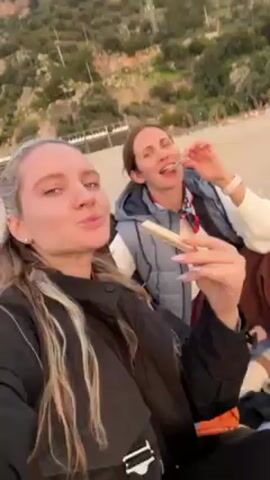 Two women are eating on a beach. The woman closest to the camera has blonde hair, a nose ring, and is wearing a black jacket. She is holding a sandwich in her hand. The other woman has brown hair, is wearing a grey vest and a white sweater, and is also eating a sandwich. In the background, a man is walking on the beach. The beach is sandy, and there is a grassy hill in the background. The sky is overcast.