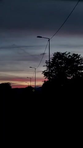 The video shows a road at dusk. The sky is a gradient of dark blue, purple, and pink. There are several streetlights along the road, which is paved. Trees and other foliage line the sides of the road, and in the distance, mountains can be seen silhouetted against the colorful sky. The camera appears to be moving, suggesting the footage was taken from a moving vehicle.