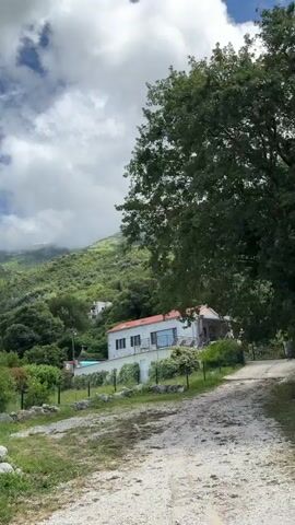 The video displays a picturesque mountainous landscape in Bratešići, Montenegro, captured from what appears to be a moving vehicle. The scene is dominated by lush green vegetation covering steep hillsides, with several houses dotted along the terrain. These houses feature stone and stucco construction, often with red-tiled roofs, and are nestled amidst trees and shrubs. Some houses have balconies and terraced gardens.

The sky is partly cloudy, with large white and grey clouds obscuring the tops of the mountains, suggesting a fresh or changing weather pattern. Patches of blue sky are visible between the clouds. The ground near the viewer is covered in wild grass and some rocky patches, indicating a rural or semi-developed area.

As the camera moves, different perspectives of the mountains and houses are revealed. A small building resembling a chapel or a small church is visible on one of the higher slopes. Power lines are also seen strung across the landscape. The overall impression is one of natural beauty and traditional architecture integrated into a rugged environment. No people or visible text are present in the video.