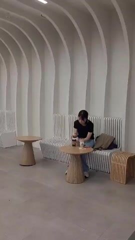 The video shows a modern, minimalist interior space with white, wavy walls and a long, continuous ceiling light. A woman in a black t-shirt and jeans sits alone at a small, light-colored wooden table in the foreground. She holds a drink and appears to be looking at her phone. To her left, a bench seat with a ribbed design is occupied by two women, also looking at their phones. Further down the hallway, more bench seating and small tables are visible. A sign displaying the number "31" is mounted on the wall in the distance. People are seen walking in the background, suggesting a public space like an airport lounge or waiting area. The overall atmosphere is calm and contemporary.