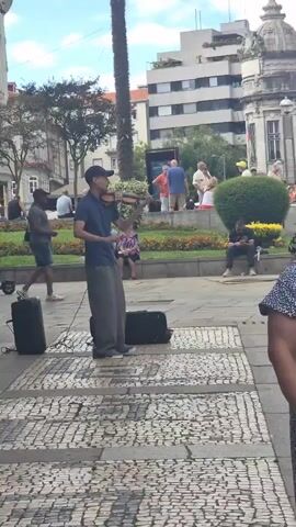 A man is playing the violin outdoors in a public square. He is wearing a blue t-shirt, a hat, and loose-fitting pants. He is standing next to a black amplifier and a black violin case. A few people are walking in the background, and others are sitting on benches. There is a palm tree and some bushes in the background, as well as buildings. The ground is paved with cobblestones.