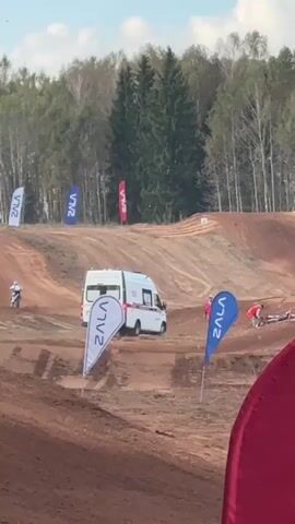 A motocross race is underway with an ambulance on standby. Several flags are visible around the dirt track. In the foreground, a person is partially visible behind a large red object. In the mid-ground, a white ambulance with red markings is parked. Several people are near the ambulance, some appear to be medical personnel. A motocross rider is seen on the track, and another rider appears to have fallen near the ambulance. In the background, trees line the top of a hill.