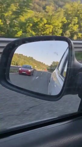 The video shows a red Tesla Model 3 driving on a highway in a mountainous area. The camera is positioned inside another vehicle, capturing the Tesla in its side mirror and then directly from behind as the Tesla drives ahead. The scene is set in a rural, natural environment with lush green trees covering the rolling hills that flank the highway. The sky appears clear and bright, suggesting it's daytime, likely late afternoon given the angle of the sunlight. The highway itself is paved with clear lane markings, indicating it's a well-maintained road.

The primary subject is the red Tesla Model 3, which is the focus of the video. The activity is the act of driving on a highway, with one vehicle following another. There are no people visible in the video, only the cars. The setting is a highway surrounded by natural, mountainous terrain.

Notable details include the reflections in the side mirror, showing the highway and other vehicles in the distance, as well as the landscape. A road sign with arrows indicating lane changes is visible on the left side of the highway at one point. The license plate of the Tesla is partially visible, reading "CA 5538 AB". The overall mood is one of peaceful travel through a scenic route. The location is confirmed as Osikovitsa, Bulgaria.